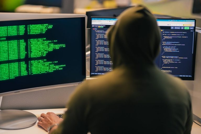 A person in a hoodie coding on dual monitors, depicting cybersecurity and hacking themes. Photo by Julio Lopez @ Pexels
