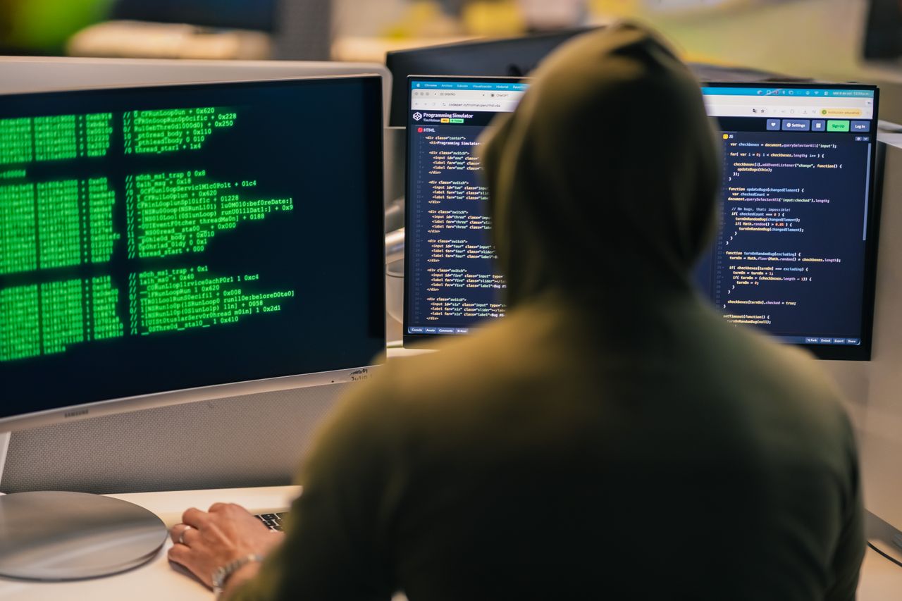 A person in a hoodie coding on dual monitors, depicting cybersecurity and hacking themes. Photo by Julio Lopez @ Pexels